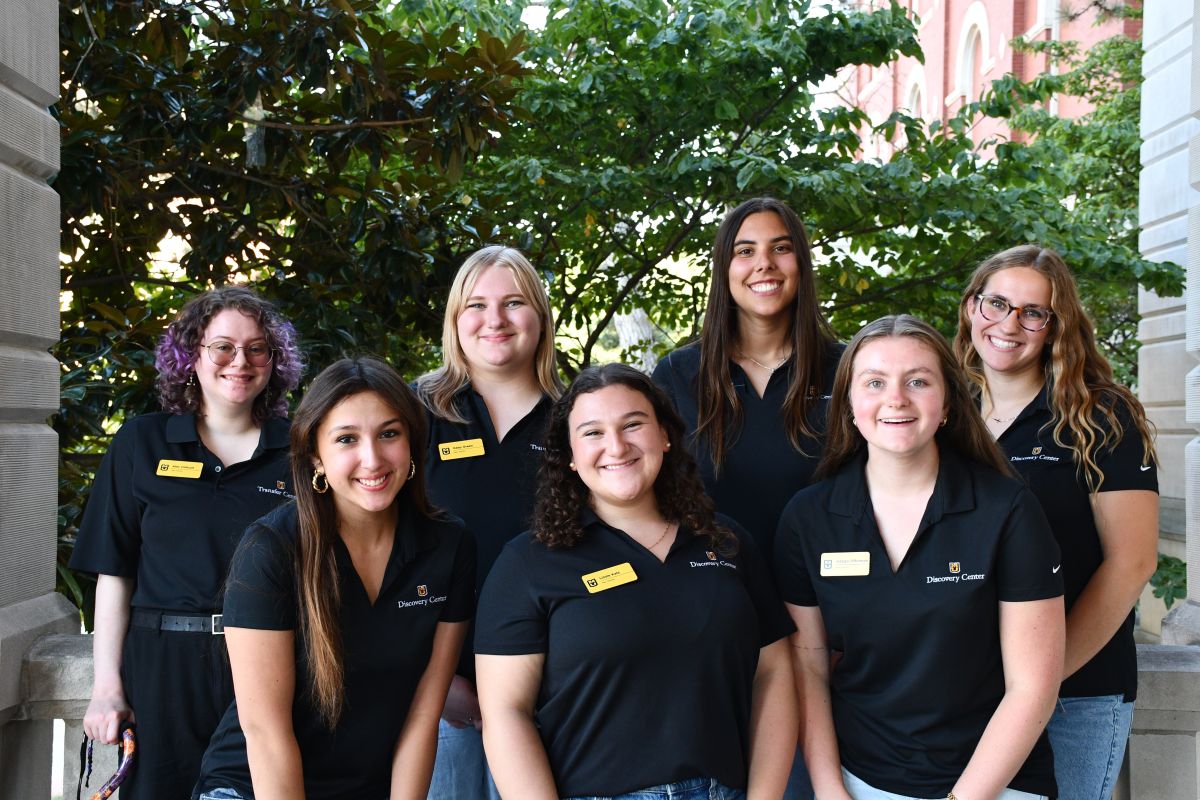 Seven Mizzou peer advisors wearing black polos stand together smiling outside Jesse Hall, with a background of green trees and bushes.
